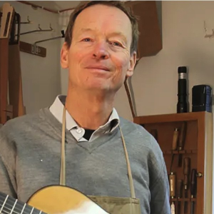 Henner Hagenlocher in a gray sweater and apron, holding a classical guitar in his workshop