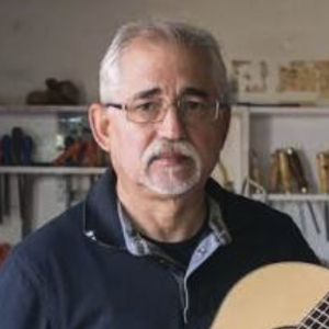 Casimiro Lozano holding a classical guitar in his workshop, with tools on the shelves behind him