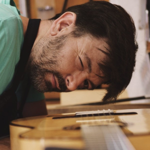 Oscar Muñoz leaning down to sight along the top of a finished guitar in his workshop