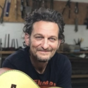Paul Aguilera smiling in his workshop with a flamenco guitar, tools hanging on the wall behind him