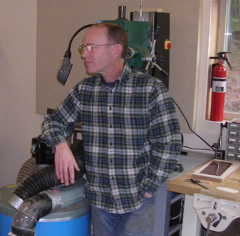 Eric Sahlin in piedi nel suo laboratorio di chitarre accanto a un aspiratore di polvere e a un banco da lavoro.