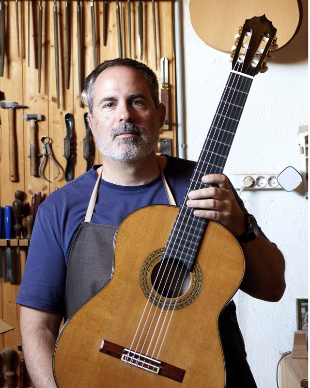 Mauricio Bellido holding a cedar-top classical guitar in front of his tool wall