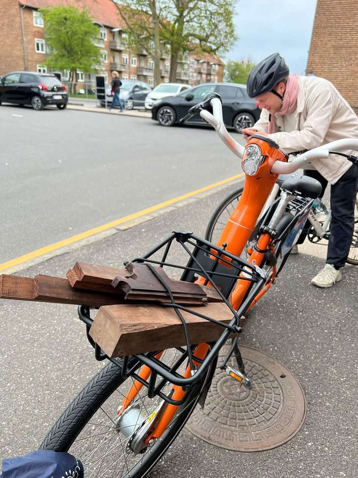 Henrik Ballowitz in a bike helmet beside an orange bicycle with rosewood boards strapped to its front rack, Copenhagen